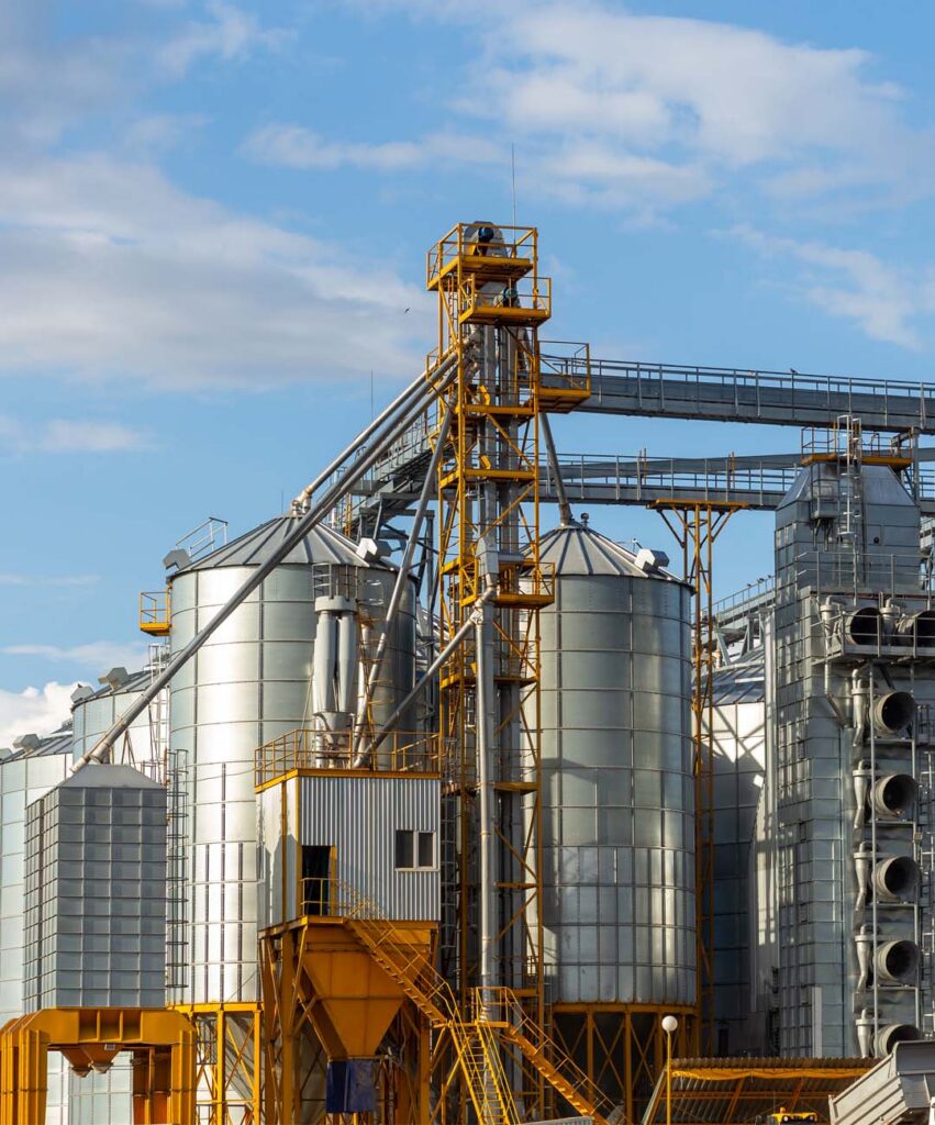 Large industrial galvanized steel grain silos and automated bucket elevator tower at a commercial agricultural storage facility.