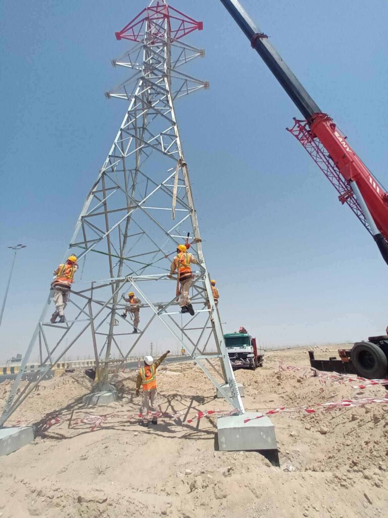 A crew of electrical workers in safety gear assembling a high-voltage metal transmission tower using a crane on an industrial construction site.