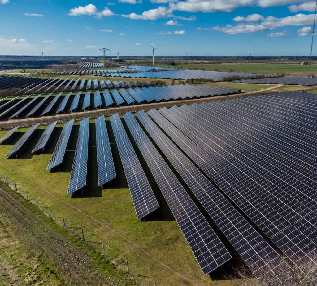 High-angle aerial view of a massive utility-scale solar farm with long rows of PV panels and wind turbines in rural green fields.
