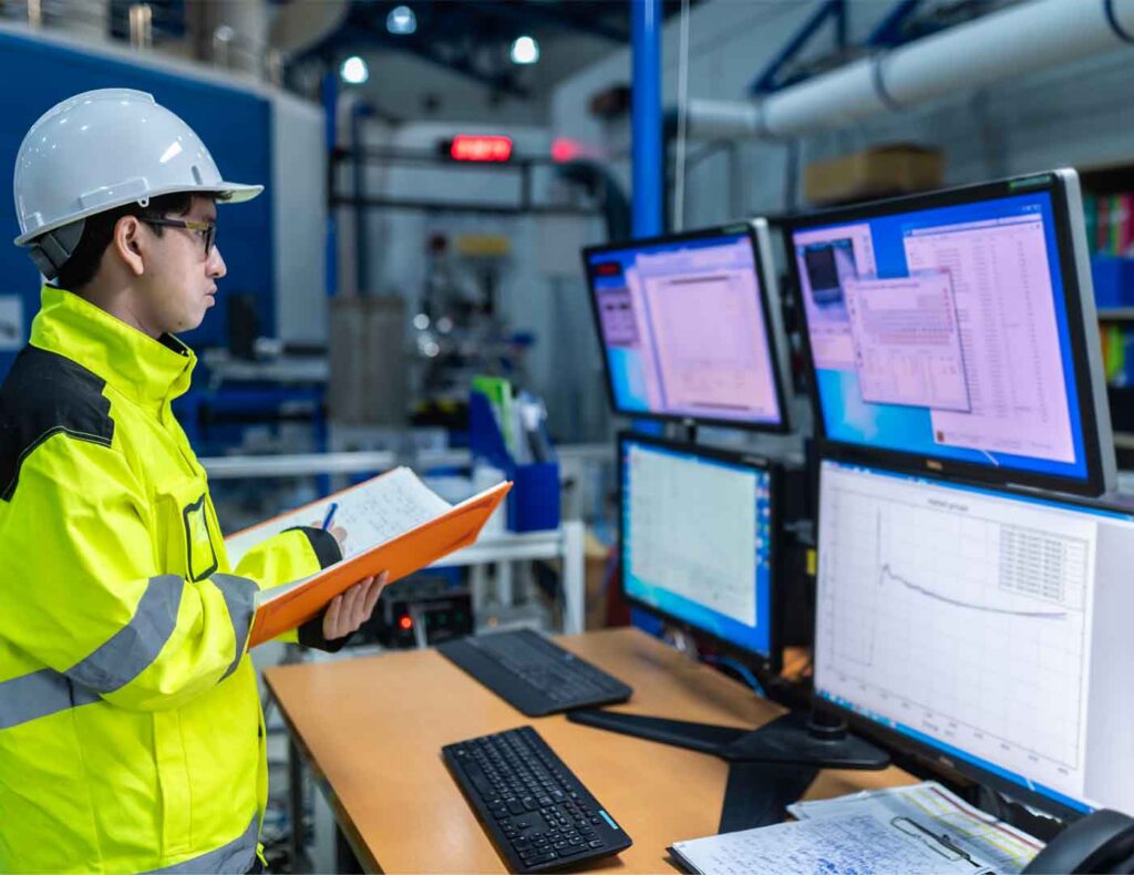 An industrial plant operator in a high-visibility jacket and hard hat monitoring complex data on multiple computer screens.