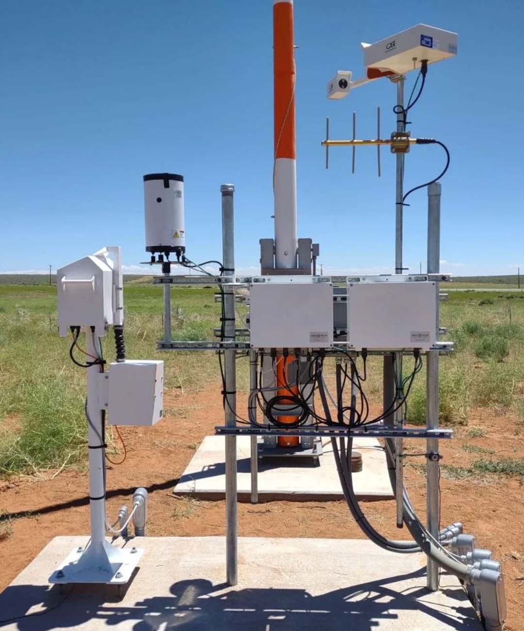 A field-deployed remote monitoring station featuring data telemetry equipment, antennas, and environmental sensors mounted on a concrete pad.