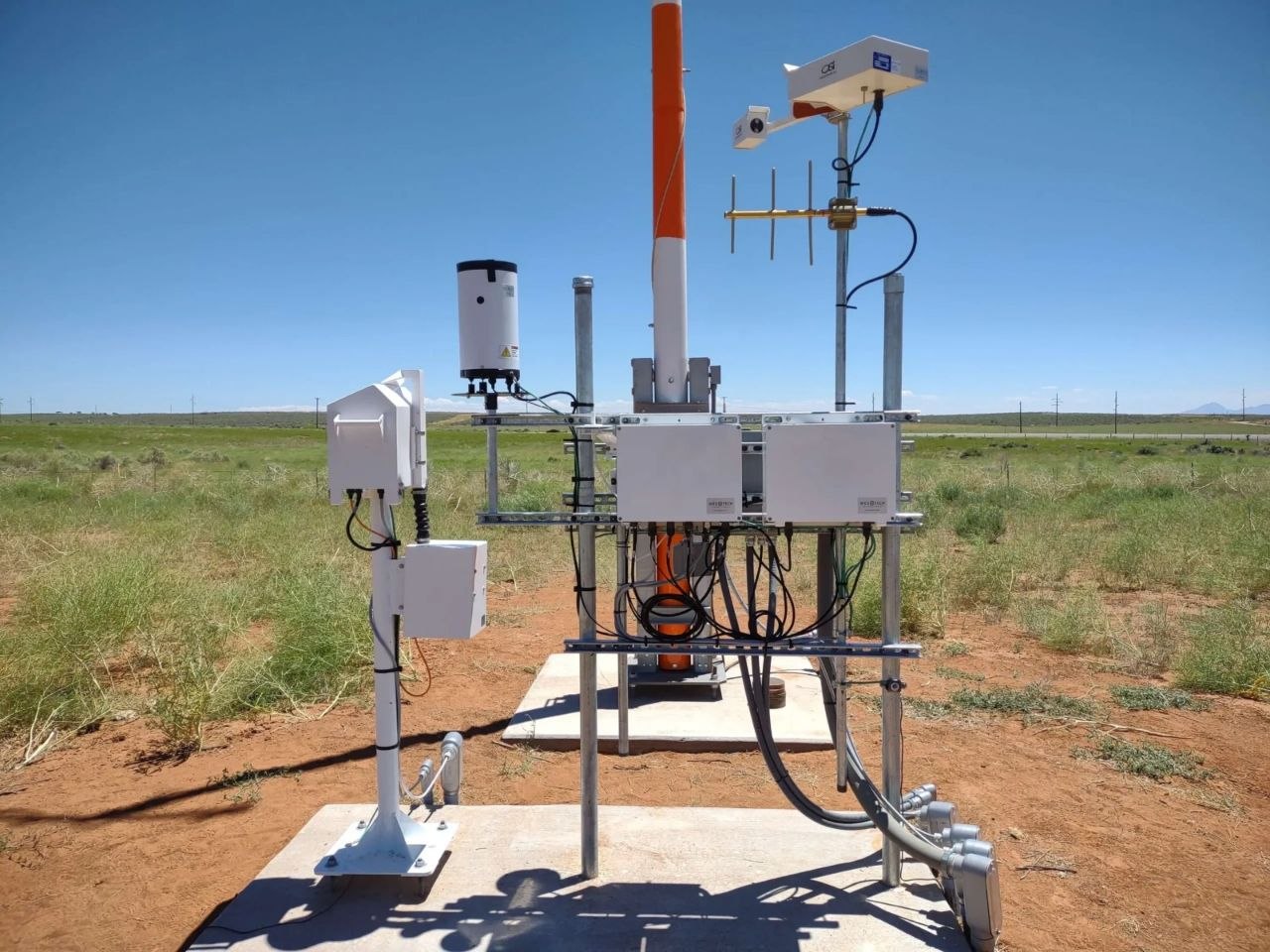 A field-deployed remote monitoring station featuring data telemetry equipment, antennas, and environmental sensors mounted on a concrete pad.